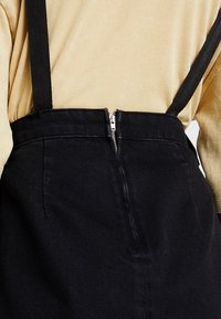 Beige långärmad skjorta instoppad i en högmidjad svart kjol med hängslen och en central silverdragkedja på baksidan.