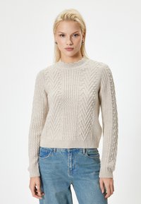 Jasno beżowy sweter z prążkowanej tkaniny, z okrągłym dekoltem, teksturowanymi wzorami oraz akcentami w postaci pomponów na rękawach i korpusie, zestawiony z niebieskimi dżinsami.