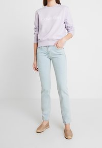 Sudadera lila con el logo blanco de "Calvin Klein", vaqueros azul claro y alpargatas beige; cuenta con puños acanalados y un ajuste relajado.