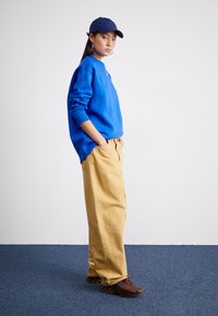 Blue sweatshirt, beige wide-leg pants, dark brown sneakers with bubble soles. Styled with a navy cap. Plain background and floor.