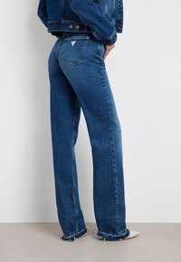 Un jean en denim bleu taille haute présente une jambe droite, un ourlet effiloché et deux poches arrière avec un logo de marque en forme de triangle.
