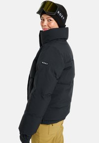 Black puffer jacket with high collar and Roxy logo on the sleeve, paired with a black beanie and ski goggles. The model has light brown hair.