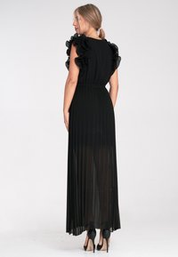 Schwarzes Maxikleid mit plissiertem Design, Rüschenschultern und tailliertem Schnitt. Durchsichtiger Stoff mit fließender Silhouette und High Heels.