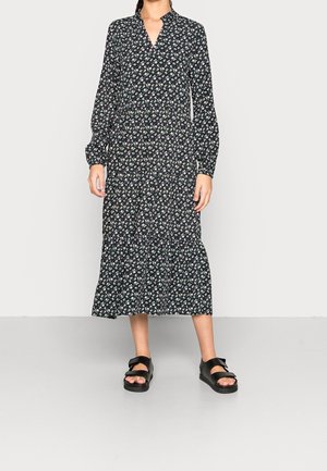 Vrouw die een zwarte midi-jurk met lange mouwen en een bloemenpatroon draagt, met een rokje in lagen, gecombineerd met zwarte open-toe sandalen met brede bandjes.