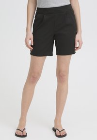 Žena v čiernych krátkych nohaviciach po kolená, šedom bezrukávniku a čiernych žabkách stojaca na bielom pozadí.