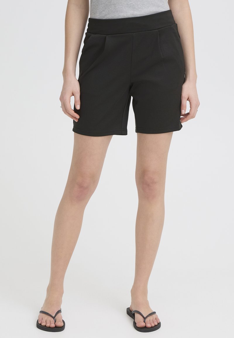 Žena v čiernych krátkych nohaviciach po kolená, šedom bezrukávniku a čiernych žabkách stojaca na bielom pozadí.