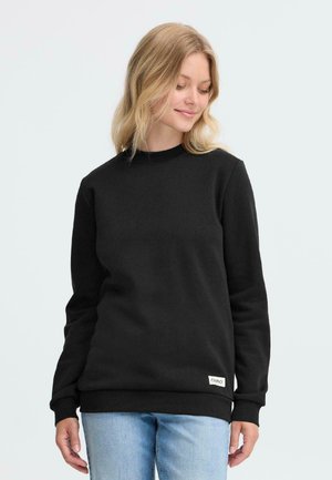 Black sweatshirt with long sleeves, round neckline, and ribbed cuffs. Features a small logo tag at the bottom hem. Worn with light blue jeans.