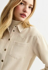 Beige corduroy shirt with a button-up front, collar, and single chest pocket. Features ribbed texture and dark buttons for accents.