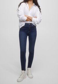 Jeans skinny de mezclilla azul de cintura alta con una textura suave, combinados con una camisa blanca de botones y zapatillas blancas de caña alta.
