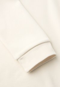 Kermanvärinen hupparin hiha ribbireunuksella sileällä kangastaustalla.