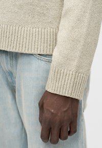 Ανοιχτό μπεζ πλεκτό πουλόβερ με ριμπ μανσέτες, φορεμένο πάνω από ανοιχτό μπλε τζιν παντελόνι, που αναδεικνύει υφή και μαλακό υλικό.