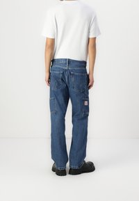 Personne portant un jean cargo en denim bleu, des chaussures noires et un t-shirt blanc uni à manches courtes, debout de dos sur un fond blanc.