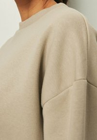 Felpa beige con collo a giro e tessuto testurizzato. Presenta spalle scese e una vestibilità rilassata, che mette in risalto linee e cuciture morbide.