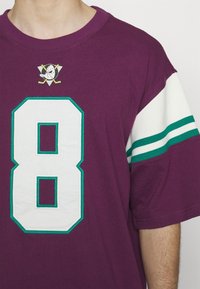 Jersey de algodón púrpura con un gran número "8" blanco con acentos en verde azulado y un logo bordado en el cuello; manga corta con rayas blancas y verdes azules.