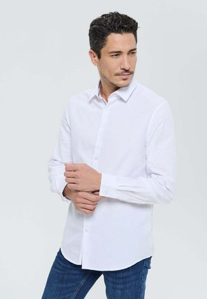Chemise blanche à manches longues avec un col classique, présentant une texture unie et des détails de col intérieur contrastants. Associée à un jean bleu.