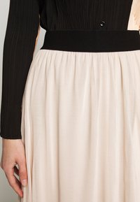 En person som bär en veckad beige kjol med en bred svart midja och en långärmad svart ribbad topp, med handen vilande vid sin sida.