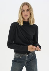 Black long-sleeve top made of a smooth fabric, featuring draped detailing at the front and a high neckline, paired with dark denim jeans.