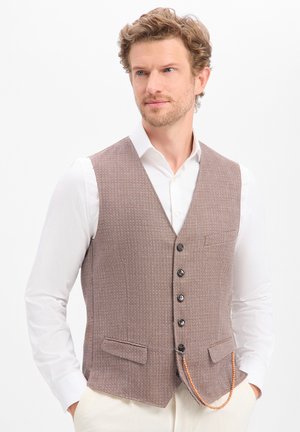 Man with curly hair wearing a textured brown buttoned vest over a white shirt, with hands in pockets and a beaded chain hanging from vest.