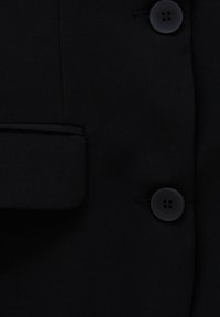 Manteau en tissu noir en gros plan montrant deux boutons noirs à quatre trous et une poche à rabat sur le côté gauche.