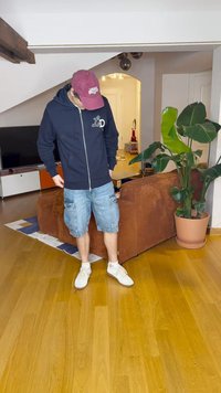 Corta-vento azul-marinho com fecho e logótipo, calções de ganga com bolsos laterais e ténis brancos, situados numa sala de estar com uma planta em vaso e móveis.