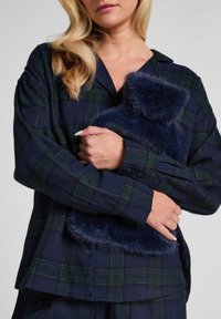 Marine ruitjes overhemd met groene lijnen en gouden accenten, gecombineerd met een donkerblauwe bontige halswarmers, vastgehouden in de armen. Zachte textuur zichtbaar.