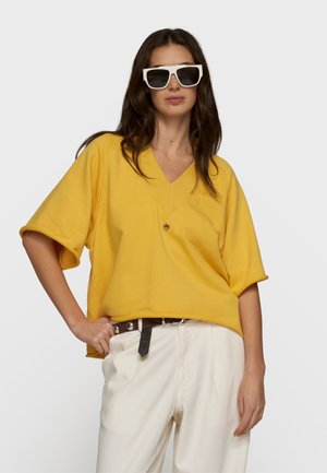 Donna che indossa una maglia gialla oversize con scollo a V, pantaloni bianchi a gamba larga, cintura marrone scuro e grandi occhiali da sole bianchi, posa con la mano sul fianco.