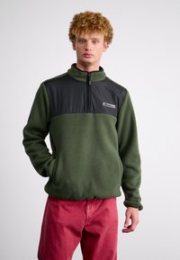 Green fleece pullover with a black panel at the chest, featuring a high collar and half-zip closure. Model wearing red pants.