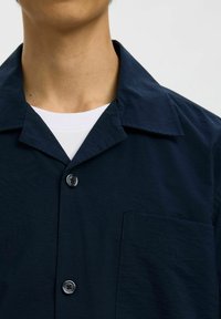 Chemise bleu marine avec col, deux boutons et une poche poitrine. Tissu lisse avec une légère texture et un design moderne.