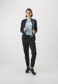 Mornarska zip-up jakna, svetlo modra majica s "PUMA" logotipom, črne jogger hlače in črne tene z zelenimi detajli. Dolgi lasje, držanje vrvi za poskočnice.