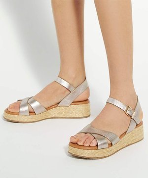 Pieds portant des sandales compensées argentées métalliques avec brides à la cheville et semelles plateforme tissées sur fond blanc.