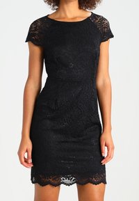 Schwarzes Spitzenkleid mit figurbetonter Silhouette, kurzen Ärmeln und wellenförmigem Saum. Die Textur zeigt durchgehend filigrane Blumenmuster.