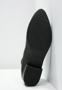 Suela de zapato de cuero negro con superficie de agarre texturizada, forma de punta ligeramente redondeada y un acabado suave. Presenta un nombre de marca estampado y una talla.