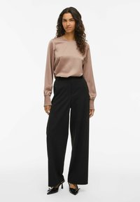 Žena v dlhom béžovom saténovom blúzke s naťahovanými manžetami, čiernych širokých nohaviciach a čiernych lodičkách na špic s mašľami.