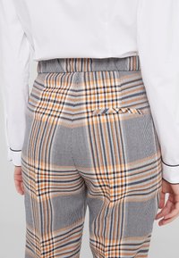 Pantalons à carreaux gris avec des rayures orange et noires, coupe structurée, et une poche passepoilée au dos, fabriqués à partir d'un tissu texturé.