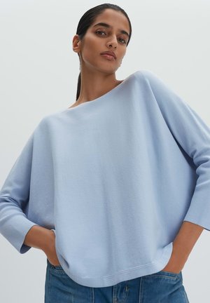 Frau mit dunklen Haaren, die einen hellblauen, locker sitzenden Pullover und blaue Jeans trägt, steht mit den Händen in den Taschen vor einem schlichten Hintergrund.