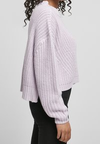 Maglione a maglia di un viola chiaro con una vestibilità ampia, polsini a coste e motivo texturizzato. Abbinato a pantaloni neri, in posizione laterale.