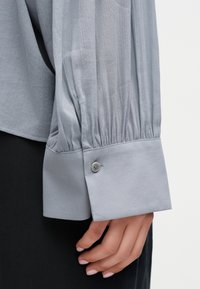 Svetlo siva majica z dolgimi rokavi z nabranim materialom nad širokim manšetom, zaprtim z enim gumbom, prikaz roke z naravnimi nohti, ki počiva ob črnih hlačah.