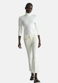 Maglione bianco a collo alto abbinato a jeans beige chiaro a zampa d'elefante con dettagli decorative sui taschini e mocassini neri lucidi.