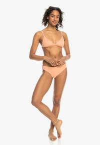 Bikiniset i ferskenfarge med ribbet tekstur; trekantet topp og vanlig passform på bunnen; tynne stropper og sømløst design. Modell står oppreist og smiler.