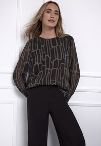 Czarna, przezroczysta bluzka z abstrakcyjnym beżowym wzorem, okrągłym dekoltem i długimi rękawami. W zestawieniu z czarnymi dopasowanymi spodniami, na neutralnym tle.