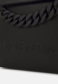 Svart textured bag med ett tjockt kedjehandtag och pr präglat varumärke "VALENTIN" på framsidan. Materialet verkar vara slitstarkt och strukturerat.