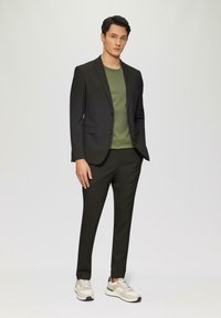 Černý oblekový sako přes zelené tričko, spojené s černými slim-fit kalhotami a béžovými teniskami s texturovaným vzorem.