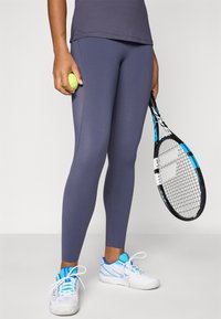 Lila sportleggings sima textúrával, fehér és kék teniszcipővel párosítva. Zöld teniszgolyót és fekete ütőt tartanak.