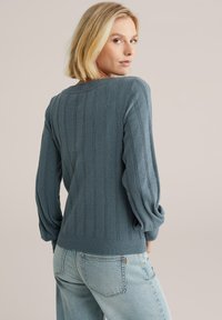 Maglione blu, lavorato a maglia con texture, design a coste, scollatura tonda e maniche lunghe a sbuffo. Abbinato a jeans chiari.