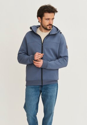 Man die de manchetten van een blauwe zip-up hoodie draagt en een spijkerbroek aan heeft, licht naar links gericht tegen een egale lichte achtergrond.