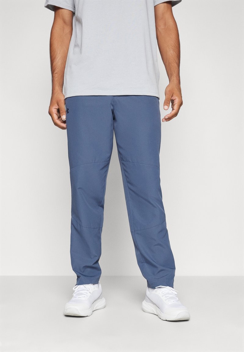 Homme portant un t-shirt gris clair, un pantalon de sport bleu et des baskets blanches, debout devant un fond clair uni.