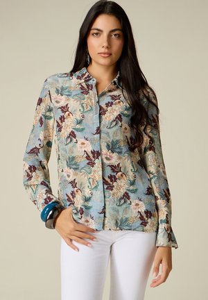 Vrouw met lang donker haar draagt een bloemetjespatroon blouse in blauw, beige en bordeaux, witte broek en grote blauwe en zwarte armbanden.