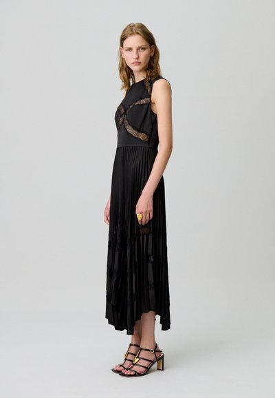 Vestido negro plisado con detalles de encaje, diseño sin mangas. La modelo lleva tacones negros con tiras, mostrando una textura de tela suave y fluida.