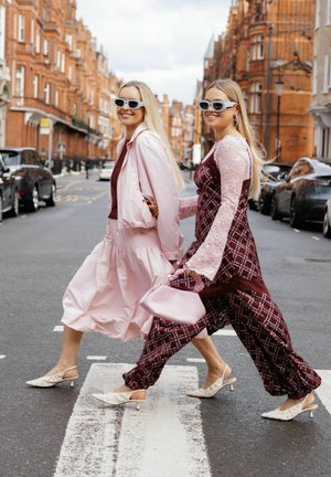 Două femei purtând ochelari de soare și rochii cu model trec strada unui oraș cu clădiri din cărămidă roșie și mașini parcate.
