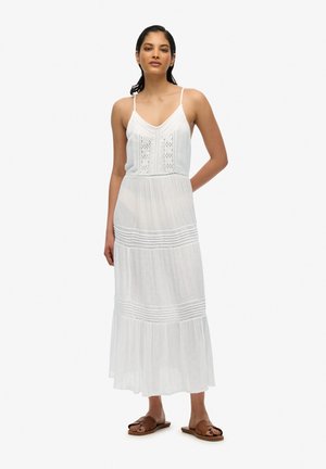 Robe maxi blanche avec un décolleté en V, des détails en dentelle au niveau du corsage, un tissu texturé et un ourlet en volants. Portée avec des sandales à brides marron.